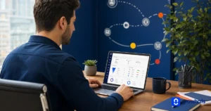 Homem trabalhando em um notebook com painel de dados e gráficos, em uma mesa de escritório; ao fundo, um painel azul com ícones de tecnologia e uma planta ao lado.